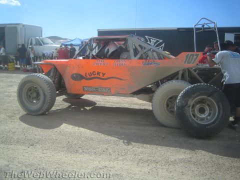 1990 Ford Bronco - orange tube-frame off-road race buggy numbered 107 and labeled "Lucky" sits in a dusty pit area beside trailers.