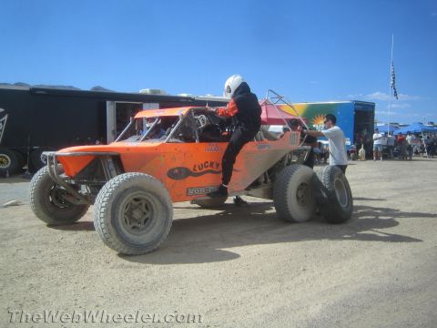 1990 Ford Bronco - driver in a red racing suit and white helmet climbs into an orange off-road race buggy under a bright desert sky.