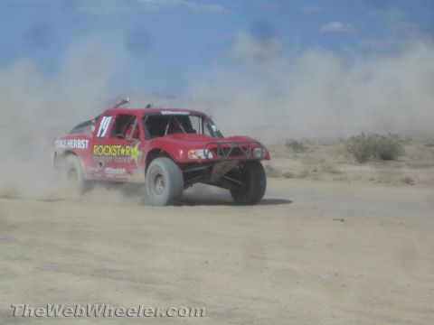 1990 Ford Bronco - red off-road race truck numbered 14 with Rockstar and Herbst livery kicks up dust racing across the desert.