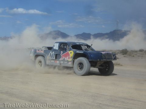 1990 Ford Bronco - blue Red Bull-liveried off-road race truck numbered 31 races through the desert raising a dust trail near distant mountains.