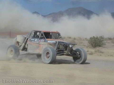 1990 Ford Bronco - orange and black tube-frame off-road race buggy speeds across the desert kicking up dust before distant mountains.
