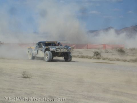 1990 Ford Bronco - black off-road race truck races past an orange course-marker fence amid heavy dust in the desert.