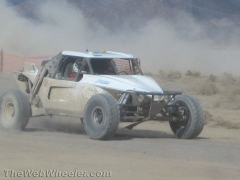 1990 Ford Bronco - white tube-frame off-road race buggy races along a dusty desert track lined with low brush.