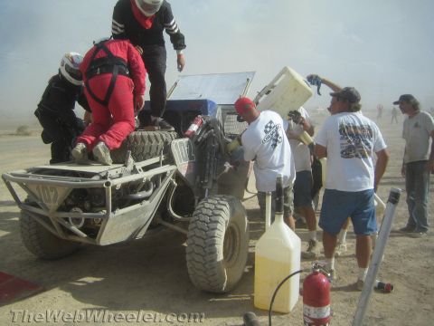 1990 Ford Bronco - pit crew refuels and services an orange off-road buggy numbered 107 with fuel jugs and a fire extinguisher at hand.