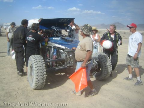 1990 Ford Bronco - Crew members and spectators gather around a blue and silver off-road buggy numbered 102 in a dusty pit area.