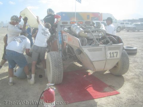 1990 Ford Bronco - pit crew refuels and services off-road buggy numbered 117 over a red mat with a fire extinguisher nearby in the dusty pits.