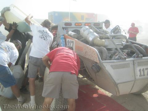 1990 Ford Bronco - Crew members lean over the rear of off-road buggy 117, exposing its engine and exhaust during a dusty pit stop.
