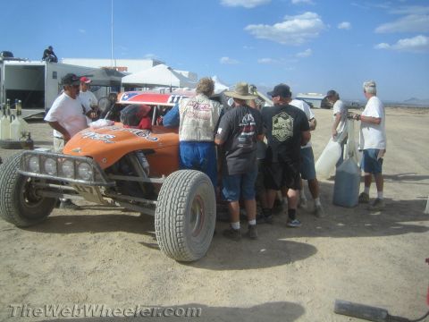 1990 Ford Bronco - group of crew members crowd around an orange off-road buggy fitted with a light bar in a dusty desert pit.
