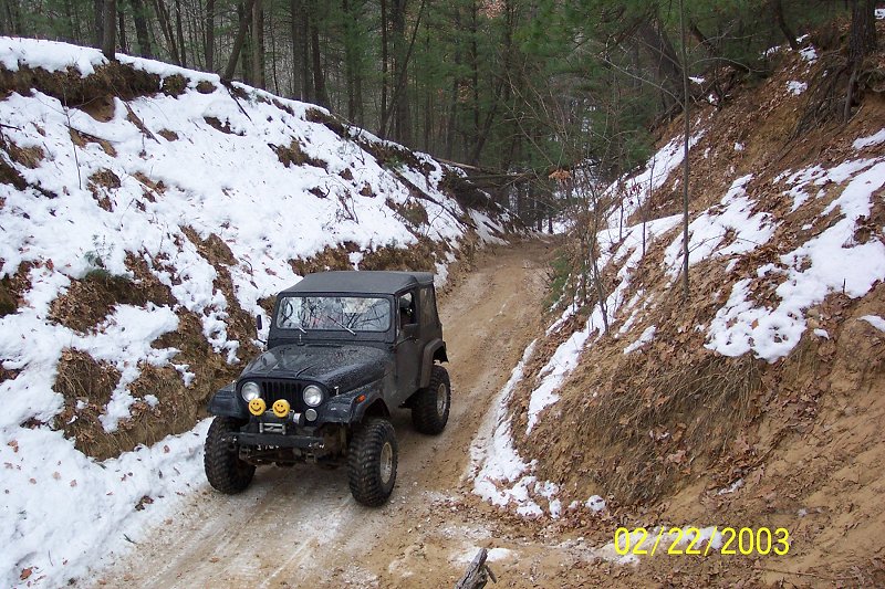 Black Jeep CJ-7 driving on a muddy, snow-dusted dirt road through a wooded area