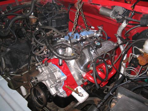 1989 Ford Bronco - Engine installation in progress, with a chain hoist supporting the motor. The red engine block and chrome valve covers are visible.