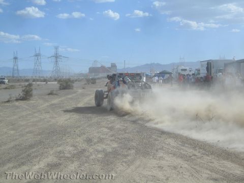 1990 Ford Bronco - off-road race vehicle trails dust as it heads toward a pit and spectator area beneath power-line towers in the desert.