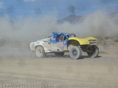 1990 Ford Bronco - blue and yellow off-road race truck races through dust in the desert beneath a tall power-line tower.
