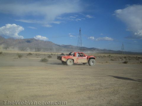 1990 Ford Bronco - red off-road race truck speeds across open desert with power-line towers and mountains in the background.