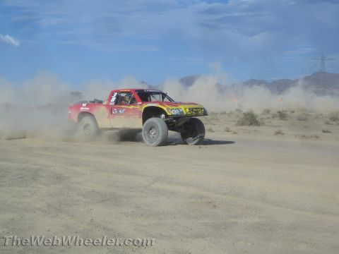 1990 Ford Bronco - red and yellow off-road race truck corners hard, throwing dust on a desert course near distant mountains.