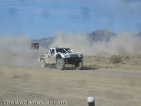 1990 Ford Bronco - white off-road race truck races through desert dust, viewed past a dust-speckled windshield.