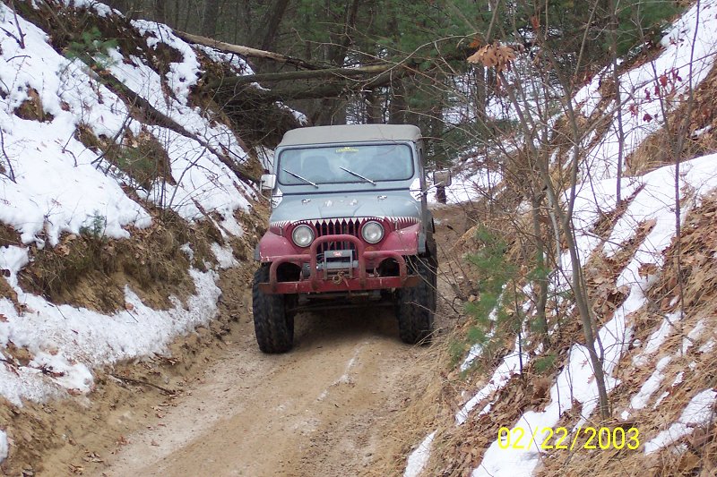 Red and gray Jeep CJ-5 with shark teeth decals on the grille, driving on a dirt path in a snowy wooded area
