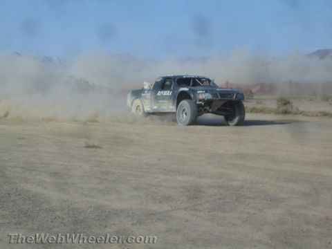 1990 Ford Bronco - dark off-road race truck with Riviera livery slides through dust on a desert course before red rock terrain.