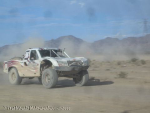 1990 Ford Bronco - white off-road race truck races through dust on a desert track with rugged mountains in the distance.