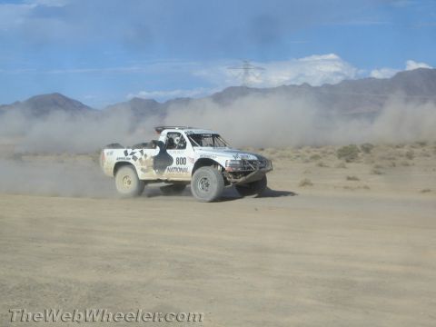 1990 Ford Bronco - white off-road race truck numbered 800 races across the desert kicking up a long dust trail near mountains.