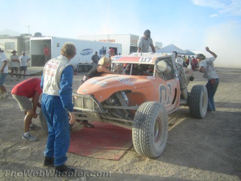 1990 Ford Bronco - Crew members in racing suits work around orange off-road buggy 117 parked on a red mat in a dusty desert pit.