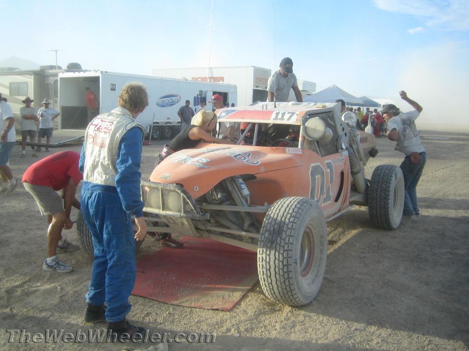 Crew around orange off-road buggy number 117 on a red mat in a dusty pit