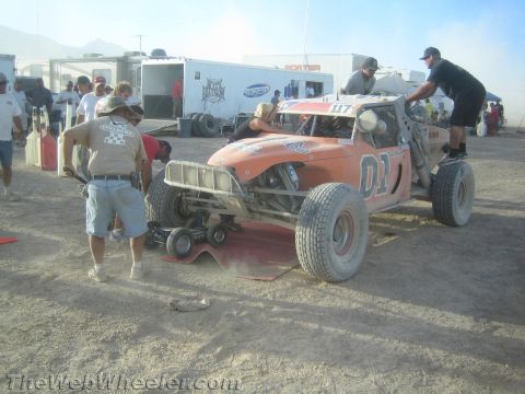 1990 Ford Bronco - Crew members work on an orange tube-frame desert race buggy marked 01 in a dusty pit area with support trailers behind.