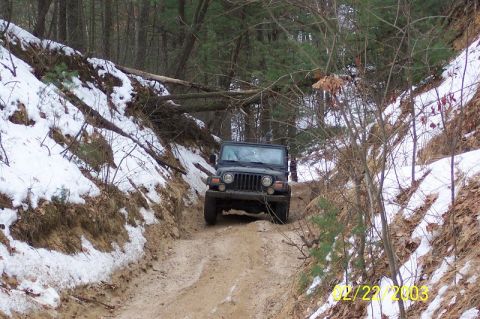 1978 Ford Bronco - Dark Jeep Wrangler with round headlights and seven-slot grille driving up a muddy snow-edged trail through pine woods.