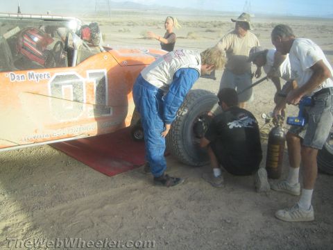 1990 Ford Bronco - pit crew swaps a wheel on an orange desert race buggy lettered Dan Myers 01 while a driver in blue racing suit looks on.