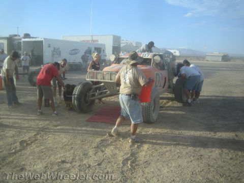 1990 Ford Bronco - group of crew members work around an orange desert race buggy in a dusty pit lined with support trailers under hazy light.