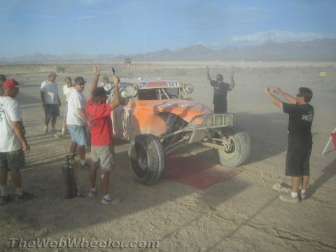 1990 Ford Bronco - Spectators raise their arms and signal beside an orange desert race buggy staged on a dusty open course with mountains behind.