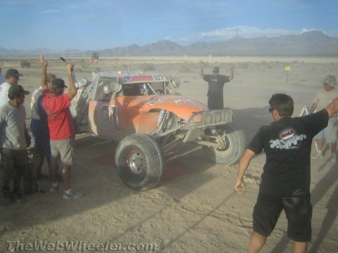 1990 Ford Bronco - orange tube-frame desert race buggy sits on a dusty staging line while crew and onlookers gesture in the hazy desert air.