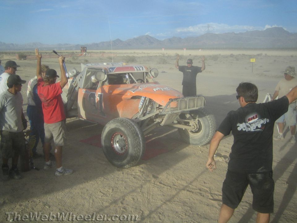 Orange tube-frame desert race buggy staged on dusty course as people gesture