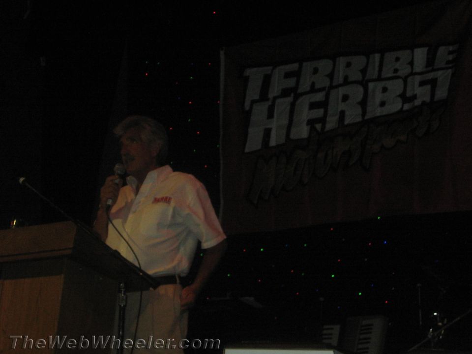 Man in white shirt speaking into a microphone at a podium before a Terrible Herbst banner