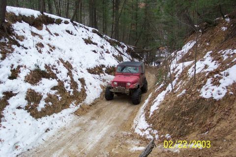 1978 Ford Bronco - A red Jeep Wrangler navigates a narrow, wooded trail with snow on the embankments.