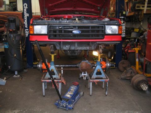 1989 Ford Bronco - Front axle assembly being worked on, with the front of a red vehicle overhead.