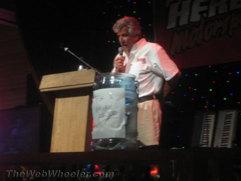 1990 Ford Bronco - gray-haired man speaks into a microphone at a podium holding a clear jar of folded papers under stage lights.