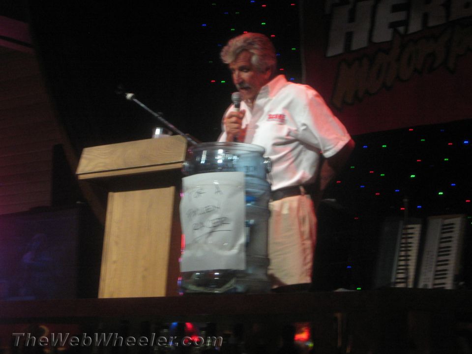 Man speaking into microphone at podium with a clear jar of papers on a dim stage