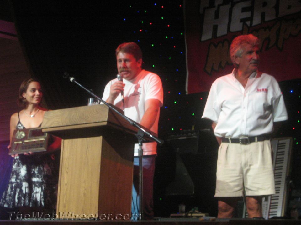 Woman holding a plaque beside two men at a stage podium with microphone