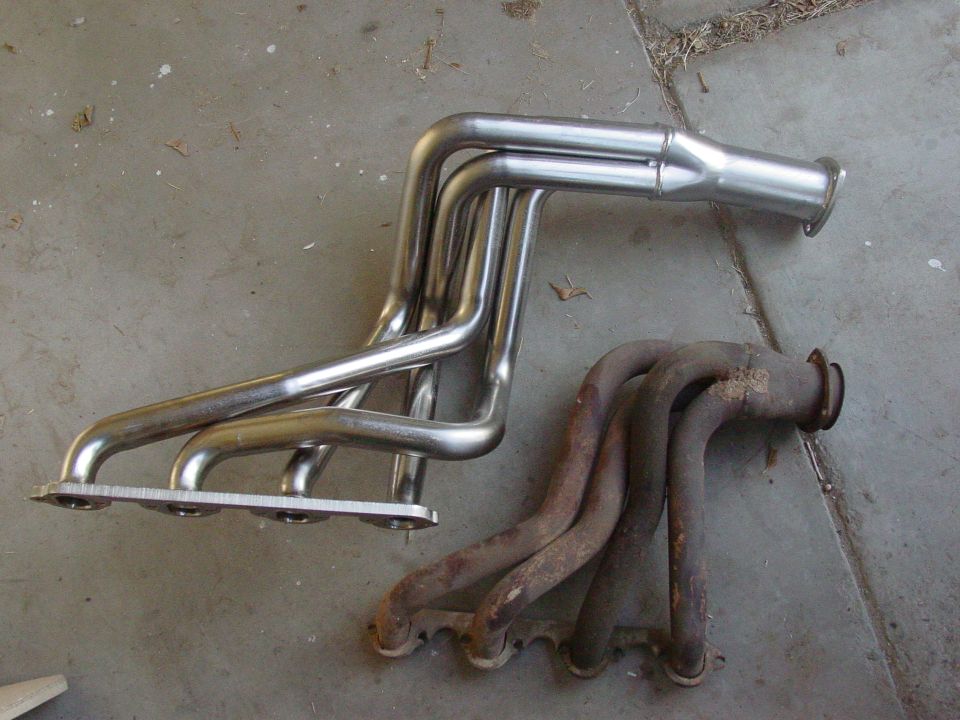 Two exhaust manifolds, one shiny and new, the other rusty and old, on a concrete surface