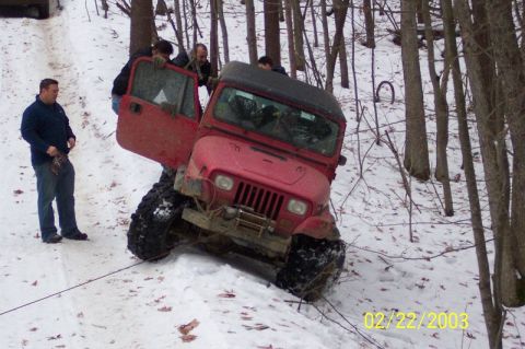 1978 Ford Bronco - A red Jeep Wrangler is stuck in a snowy wooded area, with several people attempting to assist.