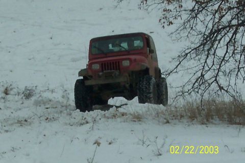 1978 Ford Bronco - Red lifted Jeep Wrangler with the seven-slot grille drives up a snow-covered hill near leafless trees.
