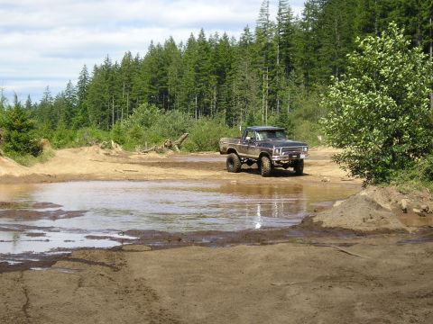 1989 Ford Bronco - A lifted Ford pickup truck navigates a muddy off-road trail with a forest in the background.