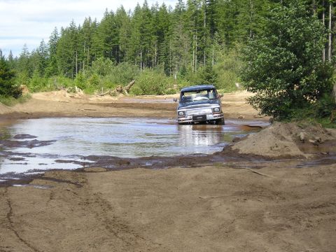 1989 Ford Bronco - A dark SUV navigates a water-filled dirt track surrounded by trees.