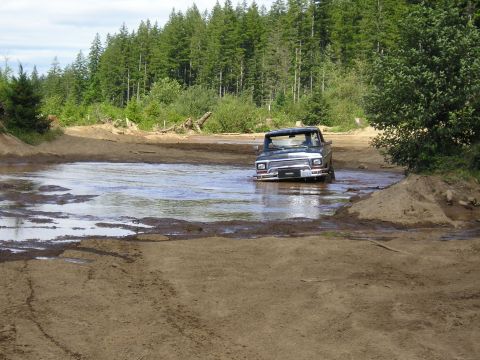 1989 Ford Bronco - A dark-colored 1980s Ford F-Series truck navigates through a muddy water obstacle in a wooded area.