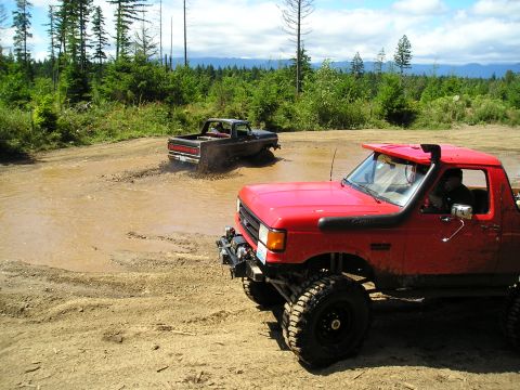 1989 Ford Bronco - Two pickup trucks navigate a muddy trail with dense forest in the background.