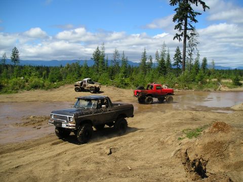 1989 Ford Bronco - Off-road vehicles navigate a challenging muddy course surrounded by dense forest and distant mountains.
