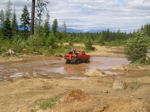1989 Ford Bronco - 1989 Ford Bronco navigating a muddy obstacle course with trees and mountains in the background.