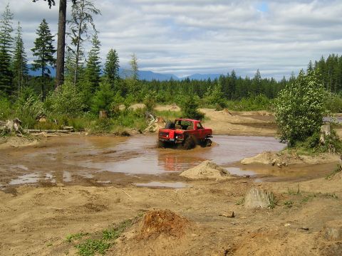 1989 Ford Bronco - A red pickup truck navigates through a muddy off-road course with trees and hills in the background.