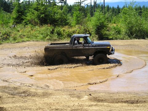 1989 Ford Bronco - A dark-colored Ford Bronco navigates a muddy trail, kicking up water and dirt.