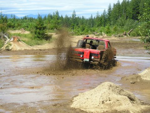 1989 Ford Bronco - A red pickup truck navigates a muddy off-road course, sending dirt and water flying.